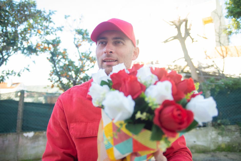 A man in a red cap and matching red jacket holds a colourful bouquet of fresh flowers, featuring red and white roses with green foliage, wrapped in patterned paper with a yellow ribbon. He is outdoors