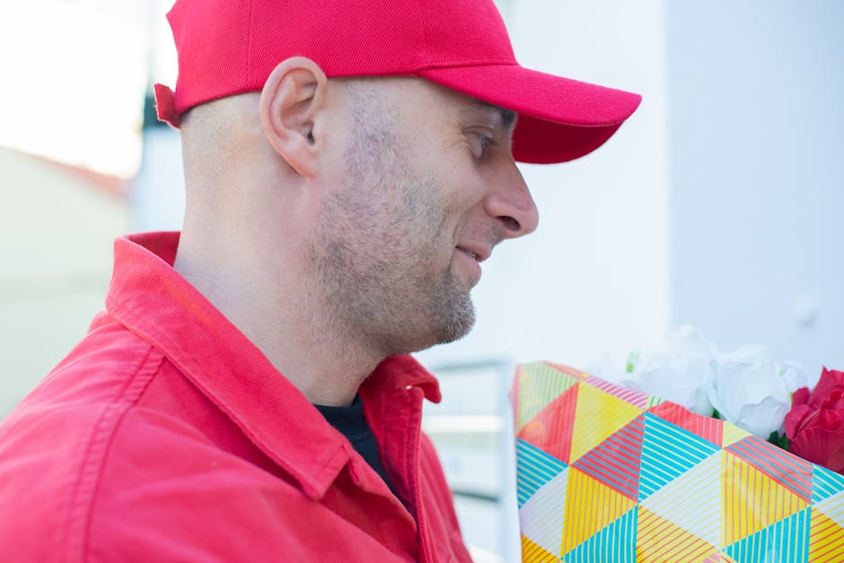A man wearing a red cap and matching red jacket is holding a vibrant, geometric-patterned gift bag filled with white and red flowers. The flowers appear fresh with lush petals and sturdy stems, arrang