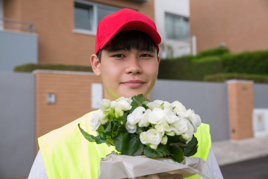 A young delivery person wearing a bright yellow high-visibility vest and a red cap is holding a bouquet of fresh white roses wrapped in white tissue paper. The roses are fully bloomed with smooth peta