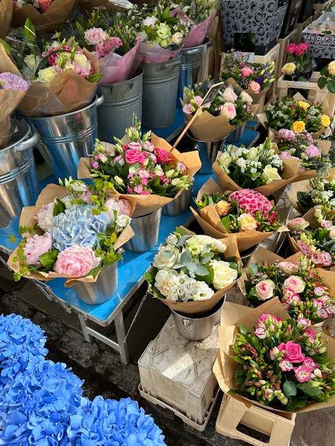 An outdoor display of fresh floral bouquets arranged in metal buckets and brown wrapping paper, featuring a variety of flowers in soft pastel shades including pink, white, and light blue. The bouquets