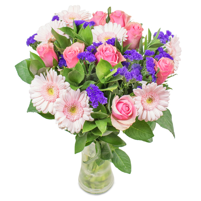 In this radiant floral arrangement, the clear glass vase reveals an expertly chosen medley of flowers that looks as though it might have been gathered from the gardens and hedgerows lining Harmondsworth Lane. Approximately seven to nine pink roses unfurl their spiral petals in stages: some tightly budded, others beginning their classic bloom, each displaying a different degree of soft-to-medium pink with a velvety sheen. Nestled between them, pale pink gerbera daisies, notable for their large, sun-like faces and lighter, ivory-tinged centers, add cheerful poise and subtle optimism. Interspersed throughout are vibrant clusters of purple statice, lending visual punctuation and textural contrast while infusing a subtle wildflower charm. At the base, dense, glossy leaves provide lush greenery, enriching the arrangement's sense of freshness and volume. The water in the vase glimmers with promise, highlighting the bouquet's vitality and suggesting it has just been delivered from a trusted Harmondsworth florist shop. Whether meant for a birthday, Mother's Day, or as a heartfelt gesture to a neighbor, this bouquet captures feelings of affection and celebration, reflecting both the care with which it was designed and the distinctive character of Harmondsworth's welcoming community.